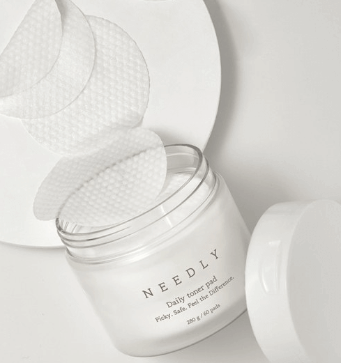 immagine suggestiva di confezione toner pads needly con coperchio aperto appoggiato in basso a destra della foto e dalla confezione escono 4 toner pads appoggiati su una superficie tipo piatto bianco